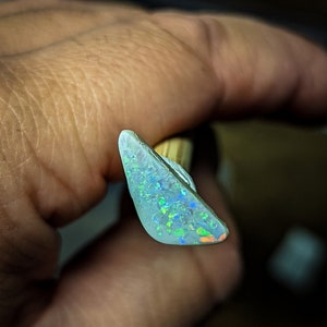 Puede incluir: Una pieza triangular de &oacute;palo blanco con destellos iridiscentes verdes, azules y naranjas.