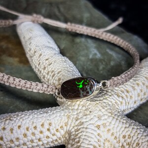 May include: A brown and green opal gemstone set in a silver pendant on a beige braided cord bracelet.