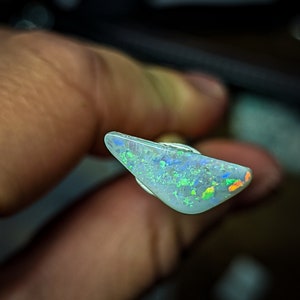Puede incluir: Una pieza triangular de &oacute;palo con una base blanca y destellos iridiscentes verdes, azules y naranjas.