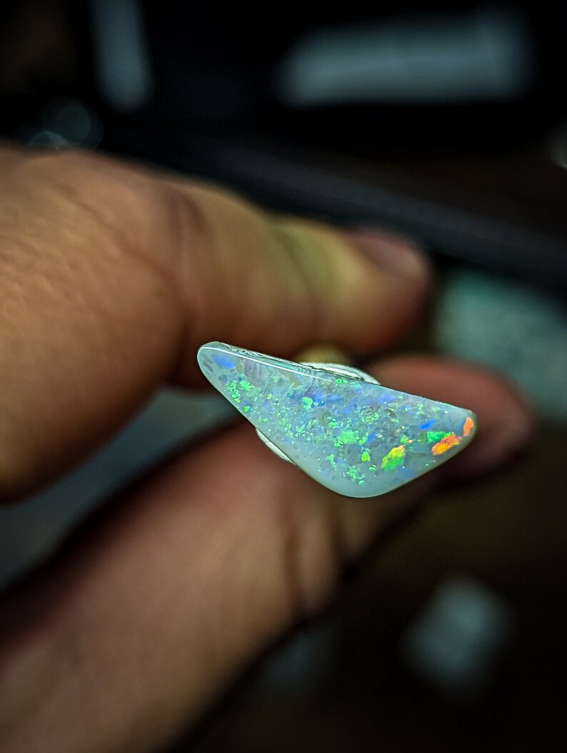 Puede incluir: Una pieza triangular de &oacute;palo con una base blanca y destellos iridiscentes verdes, azules y naranjas.
