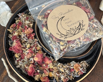 May include: A black ceramic dish with a gold rim filled with dried flowers and herbs, including rose petals and lavender. A clear plastic bag with a round label that reads "Sky Remedies New Moon Botanicals" sits on top. The background includes seashells.