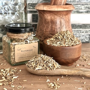 May include: A glass jar filled with dried wormwood herb, labeled "Wormwood Organic" with a pentagram charm attached to the lid. The jar is sitting on a wooden surface with a wooden mortar and pestle and a wooden spoon filled with wormwood in the foreground.