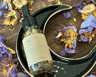 May include: A clear glass bottle with a brown label that reads "Dream Journey Intention Oil" and "Bath and Body Oil 1 fl oz 30 ml". The bottle is sitting on a black crescent moon-shaped dish with gold accents. Dried flower petals are scattered around the bottle and dish.