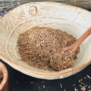 May include: A beige ceramic bowl filled with brown, dried herbs, with a wooden spoon resting inside. The bowl has a textured surface and a decorative design. A small wooden bowl is visible on the left. A bundle of dried stems is in the background.