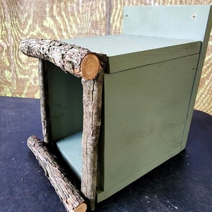 May include: A green wooden birdhouse with a square opening and a natural branch frame. The birdhouse is mounted on a wall.