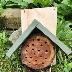May include: A wooden bee house with a green roof and a round log with holes drilled into it. The bee house is hanging from a string.