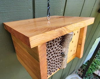 Könnte beinhalten: Ein hölzernes Bienenhaus mit einer Aufhängekette, mit verschiedenen Niströhren und einem natürlichen Ast, an dem Bienen ihre Nester bauen können.