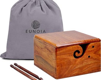 May include: A wooden yarn bowl with a lid, a gray drawstring bag with the word "EUNOIA", and two wooden crochet hooks. The yarn bowl is square-shaped with a decorative swirl cutout on the side. The wood has a warm, reddish-brown tone.