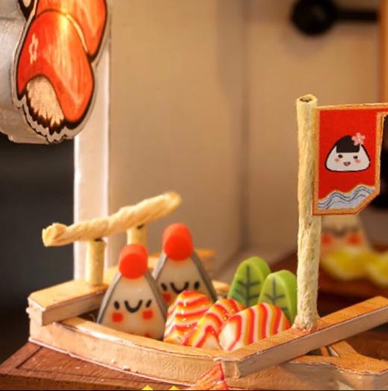 Peut inclure: Un bateau en bois miniature avec un drapeau rouge et blanc ray&eacute;. Le bateau est rempli de nourriture japonaise color&eacute;e, notamment un g&acirc;teau de poisson ray&eacute; rouge et blanc, un g&acirc;teau de poisson blanc avec un visage souriant et un g&acirc;teau de poisson blanc avec un motif ray&eacute; rouge et blanc.