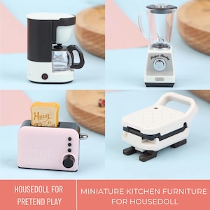 Op de afbeelding: Een set miniatuur keukenapparaten voor rollenspel, waaronder een roze broodrooster met een snee brood waarop "Hung in There" staat, een witte koffiemachine, een witte blender met de tekst "Perfect Blending" en een witte wafeltjesbak.