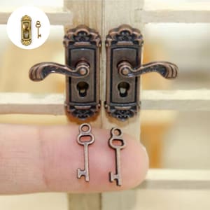 May include: Miniature bronze-colored door hardware set, including two ornate door handles and two small keys. The handles have a detailed, vintage design. The keys are held by a finger. The set is likely for a dollhouse or craft project.