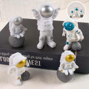 Puede incluir: Cinco figuritas de astronautas blancas con detalles dorados y azules de pie sobre bases grises con forma de luna. Los astronautas llevan trajes espaciales blancos con cascos plateados. Las figuritas están dispuestas sobre un libro negro con texto blanco.