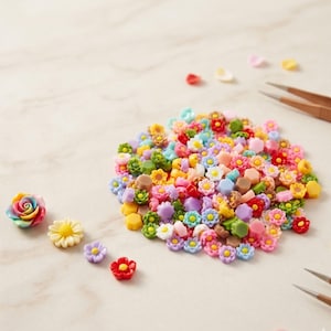 May include: A collection of colorful miniature flower embellishments in various shapes and sizes. The assortment includes roses, daisies, and other floral designs in shades of pink, yellow, blue, and green. Tweezers are visible in the background, suggesting crafting or jewelry making.