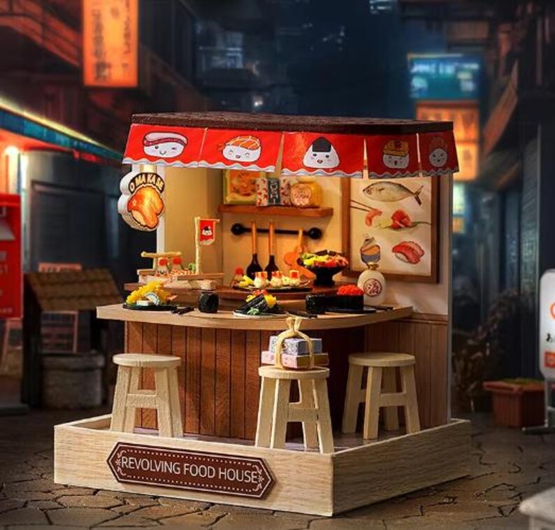 Peut inclure: Une maison de poup&eacute;e en bois miniature sur le th&egrave;me d'un restaurant tournant. La maison de poup&eacute;e est dot&eacute;e d'un auvent rouge et blanc avec une enseigne qui indique "Revolving Food House". La maison de poup&eacute;e poss&egrave;de un comptoir avec des tabourets et une vari&eacute;t&eacute; de sushis et d'autres plats japonais.