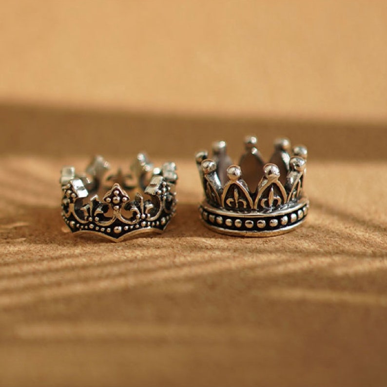 May include: Two silver crown rings with intricate details. The crowns have a pointed top and a beaded edge.