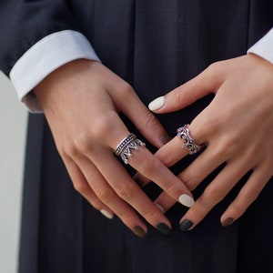 May include: Two silver rings with a crown design. The ring on the left has a more detailed design with a crown and small beads. The ring on the right has a simpler crown design. Both rings are worn on the fingers of a person's hand.