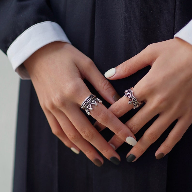 May include: Two silver rings with a crown design. The ring on the left has a more detailed design with a crown and small beads. The ring on the right has a simpler crown design. Both rings are worn on the fingers of a person's hand.