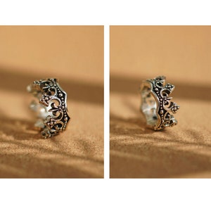 May include: A silver crown ring with intricate details. The ring is on a light brown surface.