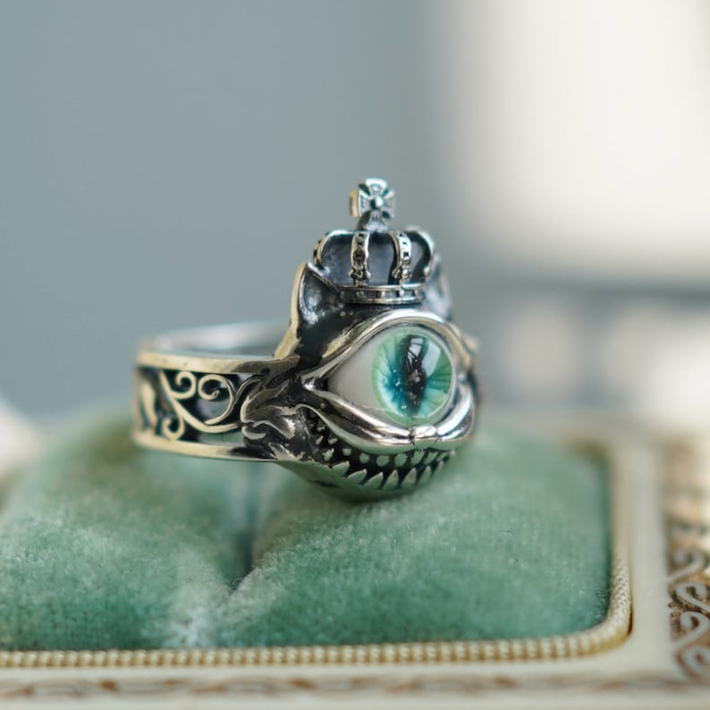 Pu&ograve; includere: Un anello d'argento con un designo dettagliato che presenta una corona, un occhio con iride verde e una bocca stilizzata. L'anello &egrave; su una superficie di velluto verde.