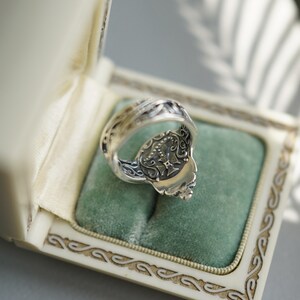 Pu&ograve; includere: Un anello d'argento con dettagli intricati e un design sul retro. L'anello &egrave; in una scatola bianca con un rivestimento in velluto verde.