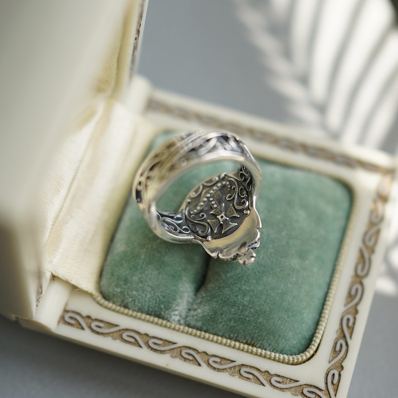 Pu&ograve; includere: Un anello d'argento con dettagli intricati e un design sul retro. L'anello &egrave; in una scatola bianca con un rivestimento in velluto verde.