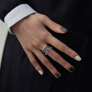 May include: A silver crown ring with intricate details. The ring is worn on the middle finger of a hand with painted nails.