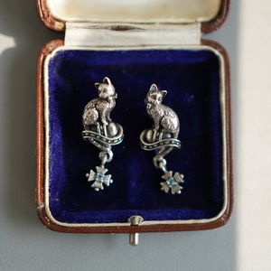 Op de afbeelding: Een paar zilveren kattenoorbellen met een blauw stenen detail. De oorbellen zitten in een blauwe fluwelen juwelendoos.