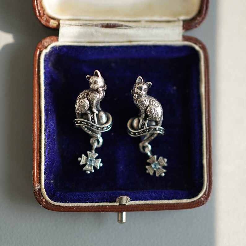 Op de afbeelding: Een paar zilveren kattenoorbellen met een blauw stenen detail. De oorbellen zitten in een blauwe fluwelen juwelendoos.