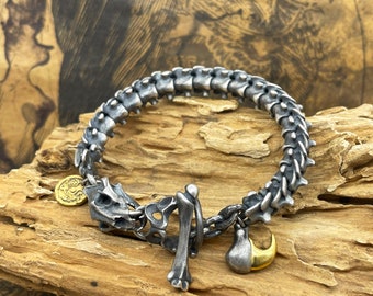 May include: A silver bracelet with a skeletal design, featuring a skull, bone, and a small charm. The bracelet is made of metal and has a chain link closure.