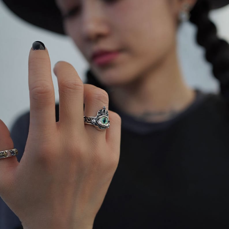 Pu&ograve; includere: Un anello d'argento con un occhio verde e una corona in cima. L'anello &egrave; indossato al dito di una persona.