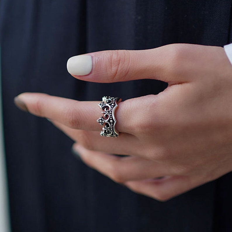 May include: A silver crown ring with intricate details. The ring is worn on a finger.