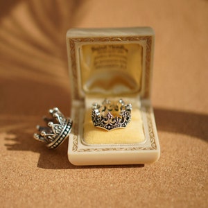 May include: A silver crown ring in a velvet-lined box. The ring is detailed with intricate designs and sits on a yellow cushion.