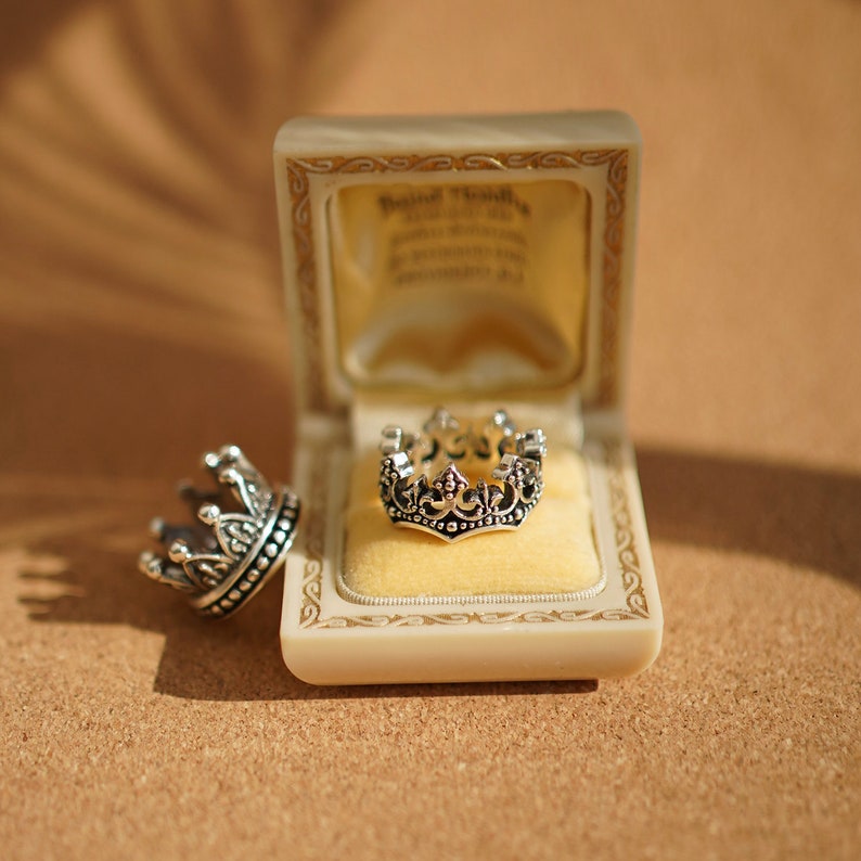 May include: A silver crown ring in a velvet-lined box. The ring is detailed with intricate designs and sits on a yellow cushion.