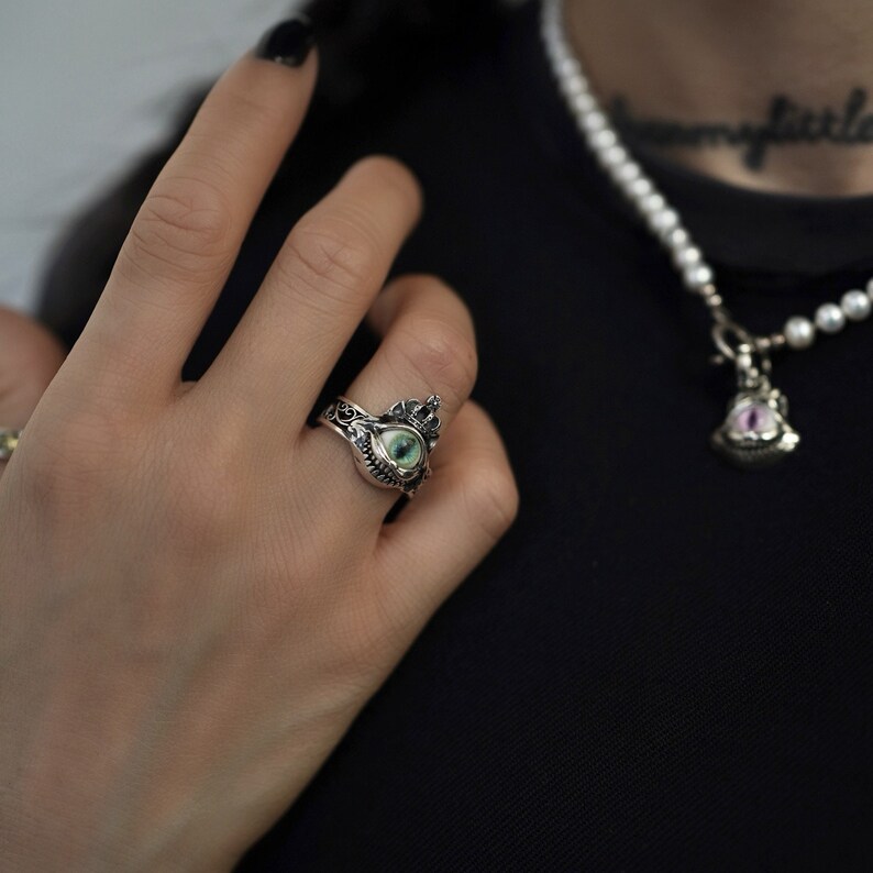 Pu&ograve; includere: Un anello d'argento con un occhio verde e un design a corona. L'anello &egrave; indossato al dito di una persona.