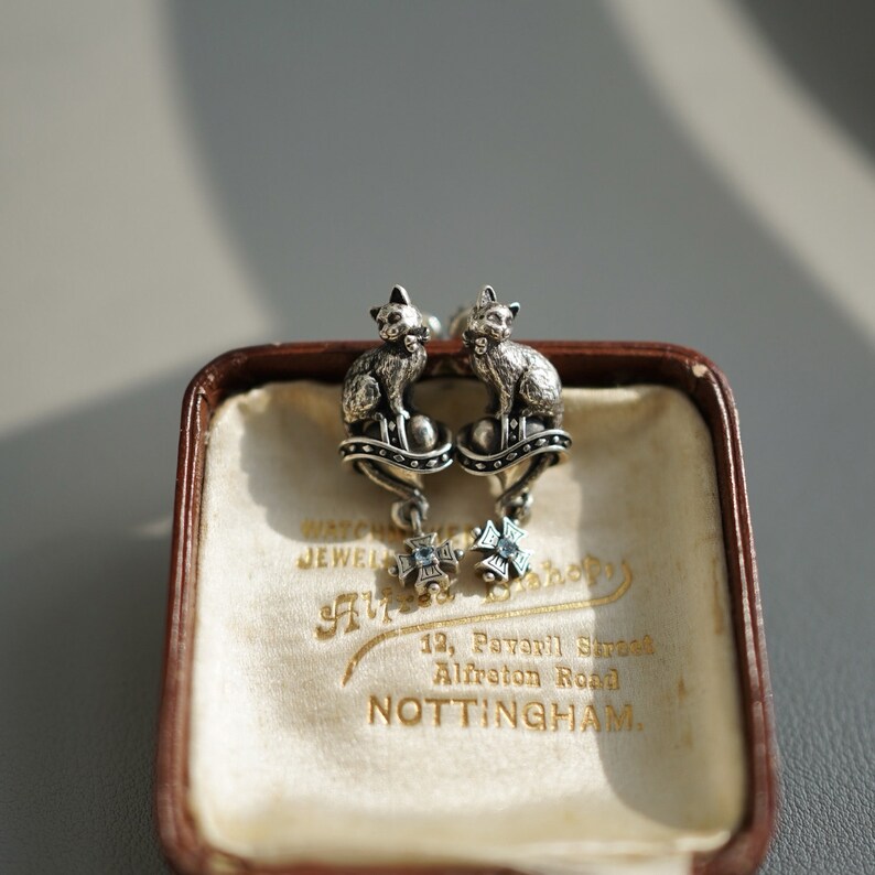 Op de afbeelding: Een paar zilveren kattenoorbellen met blauwe edelsteenaccenten. De oorbellen worden gepresenteerd in een kleine, bruine fluwelen doos met een wit label waarop staat "West End Jewellery Shop, 12, Pavell Street, Alfreton Road, NOTTINGHAM."