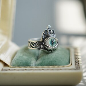 Pu&ograve; includere: Un anello d'argento con un designo dettagliato che presenta una corona, un grande occhio con un'iride verde e un ghigno dentato. L'anello &egrave; esposto in una scatola foderata di velluto.