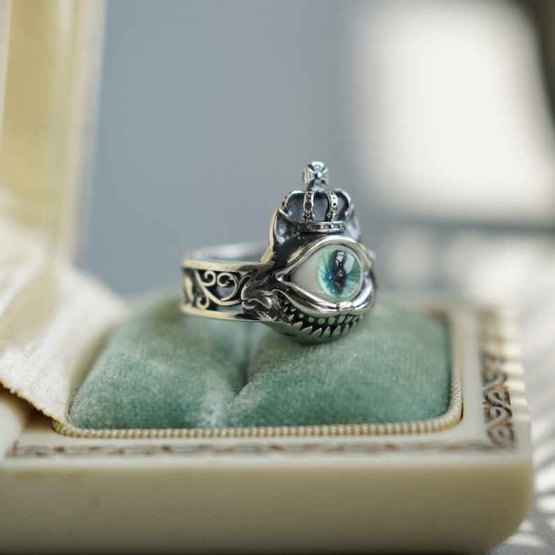 Pu&ograve; includere: Un anello d'argento con un designo dettagliato che presenta una corona, un grande occhio con un'iride verde e un ghigno dentato. L'anello &egrave; esposto in una scatola foderata di velluto.