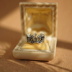 May include: A silver crown ring in a white velvet-lined box. The box is made of white plastic and has a gold trim.
