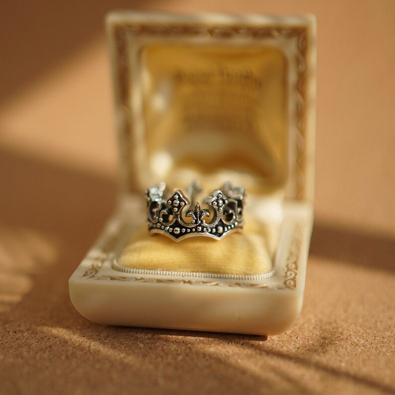 May include: A silver crown ring in a white velvet-lined box. The box is made of white plastic and has a gold trim.