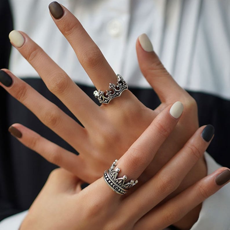 May include: Two silver crown rings. The rings are worn on the index and middle fingers of a hand. The rings are detailed with intricate designs.