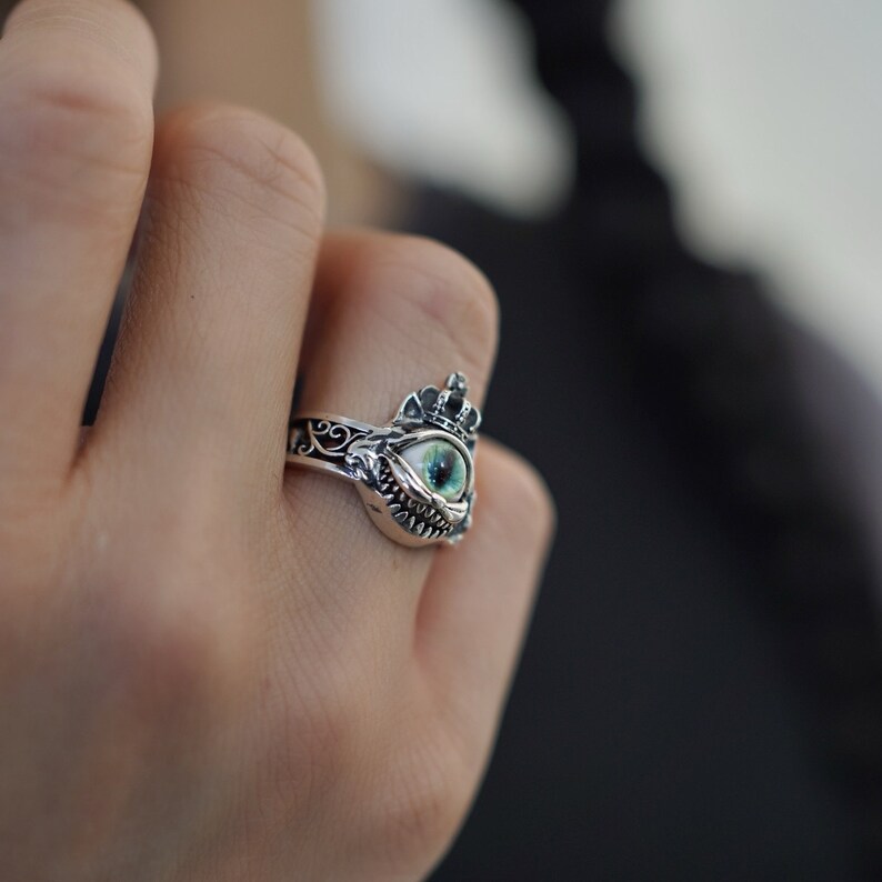 Pu&ograve; includere: Un anello d'argento con un designo dettagliato che presenta un occhio verde con una corona in cima. L'occhio &egrave; circondato da una cornice d'argento con un designo dettagliato.