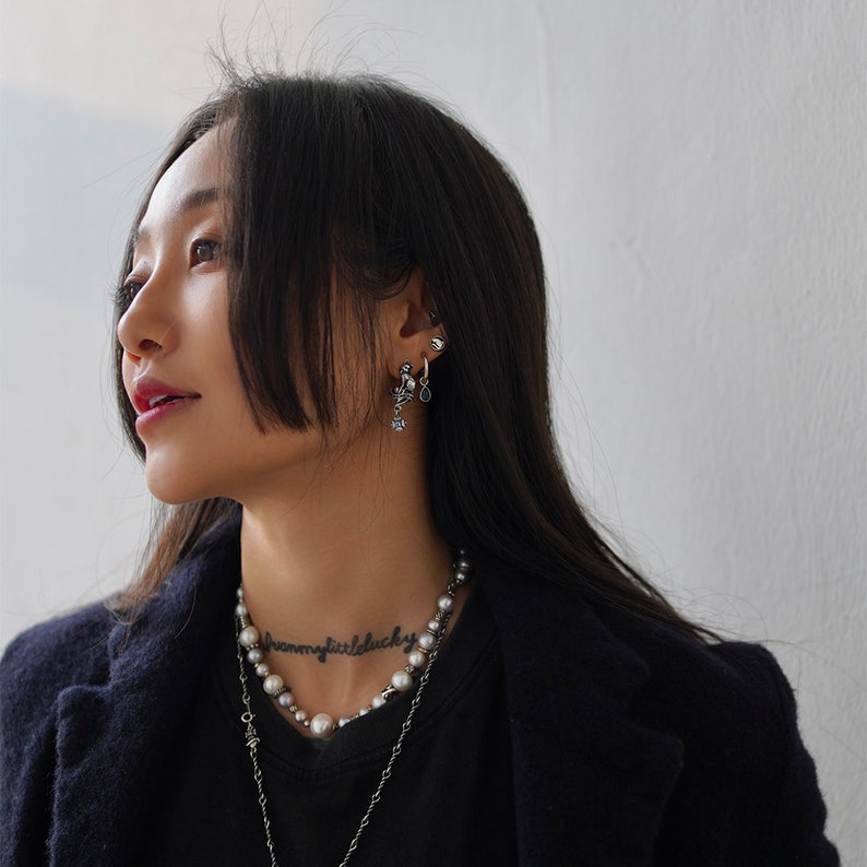 Op de afbeelding: Een vrouw met lang zwart haar draagt een zwarte blazer, een zwart shirt en een zilveren ketting met een parelketting. Ze heeft een tatoeage op haar nek die "dream a little lucky" zegt. Ze draagt meerdere oorbellen in haar linker oor.