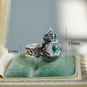 Pu&ograve; includere: Anello d'argento con un designo dettagliato che presenta una corona, un occhio verde e un ghigno dentato. L'anello &egrave; alto 17 mm e largo 5 mm.
