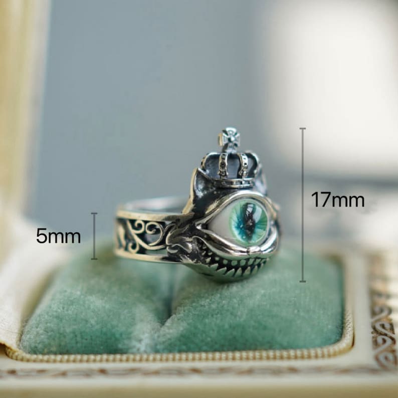 Pu&ograve; includere: Anello d'argento con un designo dettagliato che presenta una corona, un occhio verde e un ghigno dentato. L'anello &egrave; alto 17 mm e largo 5 mm.