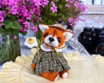 Puede incluir: Una muñeca de zorro hecha a mano con pelaje naranja, que lleva un vestido a cuadros verdes, un sombrero de paja y un cuello blanco. La muñeca sostiene una margarita blanca y tiene un velo. El fondo incluye flores rosas y tela amarilla.