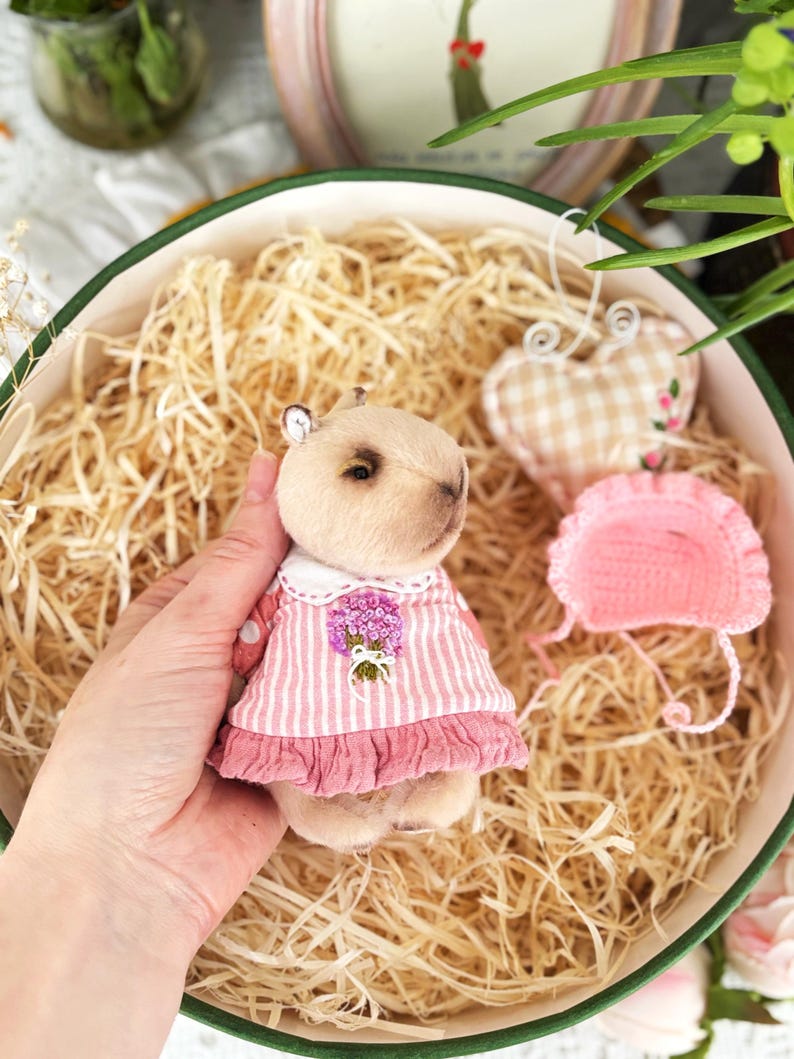 K&ouml;nnte beinhalten: Ein handgefertigtes Pl&uuml;sch-Wasserschwein, das ein rosa-wei&szlig; gestreiftes Kleid mit wei&szlig;em Kragen und floraler Verzierung tr&auml;gt. Das Wasserschwein liegt in einer dekorativen Schale mit Stroh, zusammen mit einem herzf&ouml;rmigen Ornament.