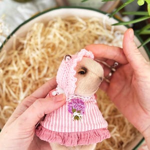 K&ouml;nnte beinhalten: Ein kleines Stofftier, m&ouml;glicherweise ein B&auml;r, tr&auml;gt ein rosa gestreiftes Kleid mit R&uuml;schensaum und passender Haube. Das Kleid hat einen gestickten Blumenstrau&szlig;. Das Spielzeug wird in den H&auml;nden einer Person gehalten.