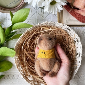 K&ouml;nnte beinhalten: Ein handgefertigter brauner Teddyb&auml;r mit gelbem Kragen und schwarzen Punkten, eingebettet in einem geflochtenen Korb. Der B&auml;r ist von trockenem Stroh umgeben und wird von einer Person gehalten. Wei&szlig;e G&auml;nsebl&uuml;mchen und gr&uuml;ne Bl&auml;tter sind im Hintergrund.