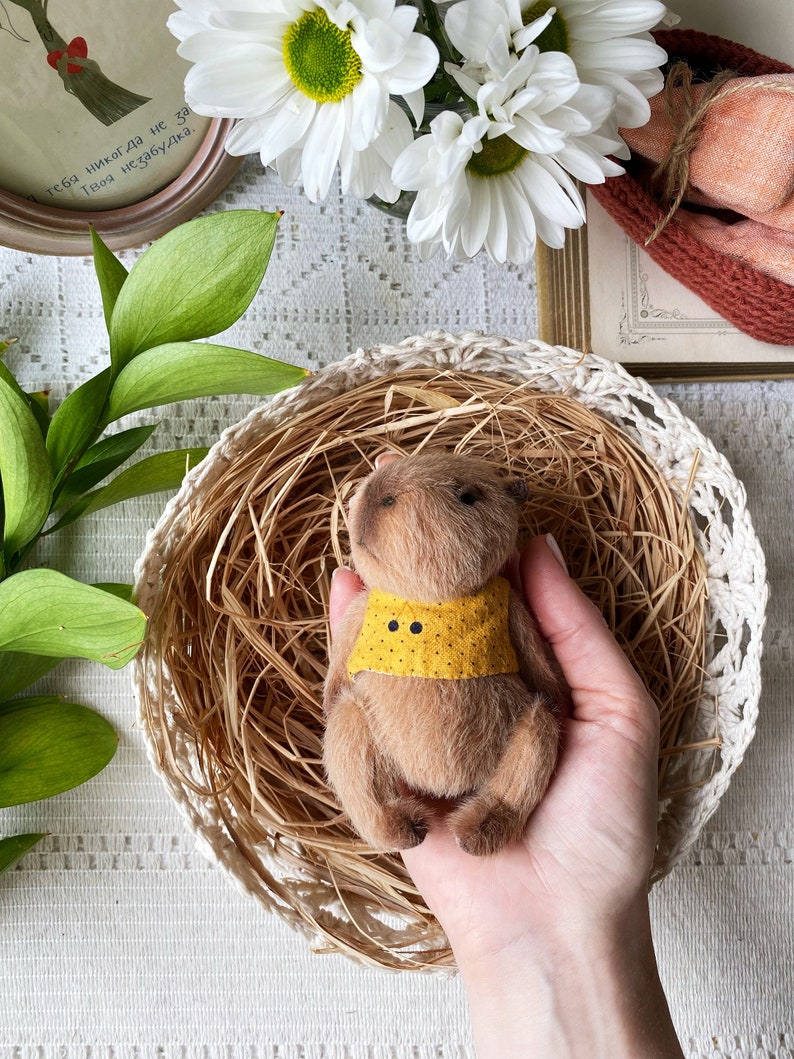 K&ouml;nnte beinhalten: Ein handgefertigter brauner Teddyb&auml;r mit gelbem Kragen und schwarzen Punkten, eingebettet in einem geflochtenen Korb. Der B&auml;r ist von trockenem Stroh umgeben und wird von einer Person gehalten. Wei&szlig;e G&auml;nsebl&uuml;mchen und gr&uuml;ne Bl&auml;tter sind im Hintergrund.