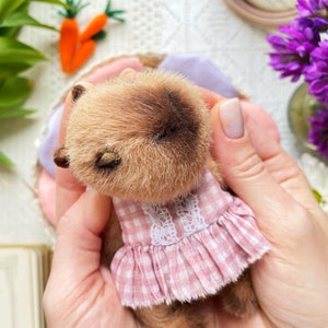 K&ouml;nnte beinhalten: Ein handgefertigter kleiner Teddyb&auml;r mit braunem Fell und einem rosa Vichy-Kleid mit wei&szlig;er Spitzenborte. Der B&auml;r hat geschlossene Augen und wird in H&auml;nden gehalten. Im Hintergrund sind Karotten und lila Blumen zu sehen.